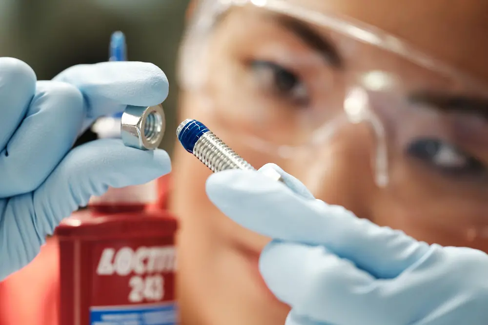 A woman's face in the background. In the front her hands with blue gloves holding a screw with blue Threadlocker.