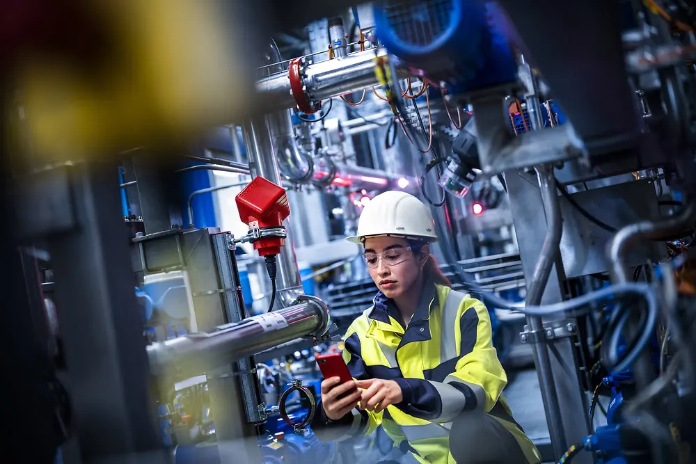 A female worker is standing in a production site checking on her mobile device the status of the equipment.