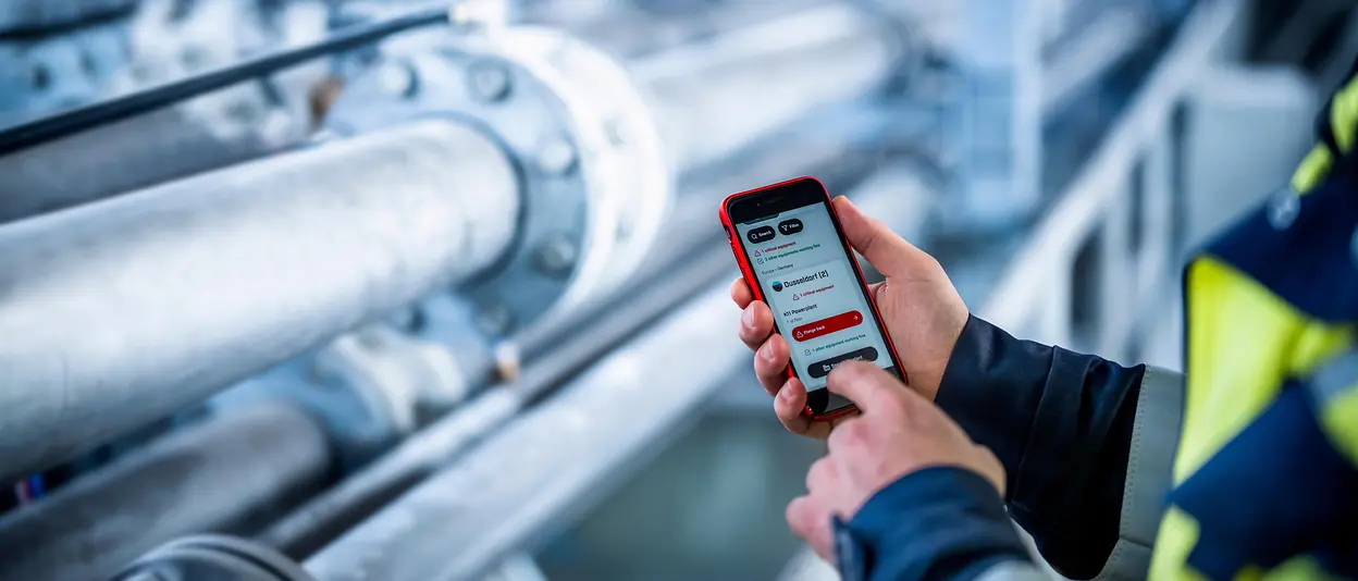 A production worker is holding a smart phone standing in front of equipment to do maintenance.