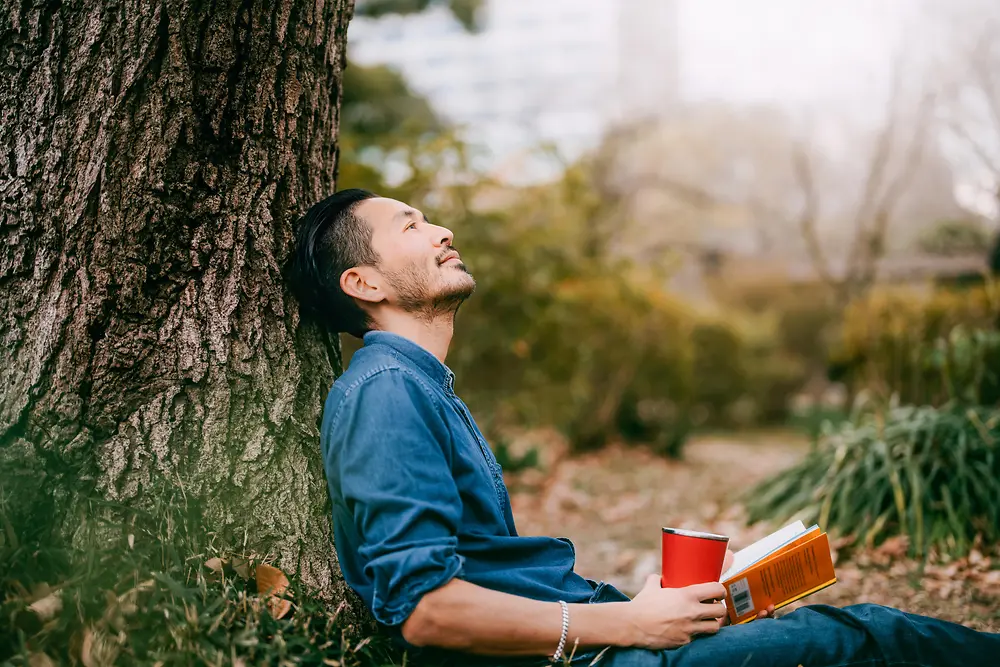 A man with a book leaning against a tree