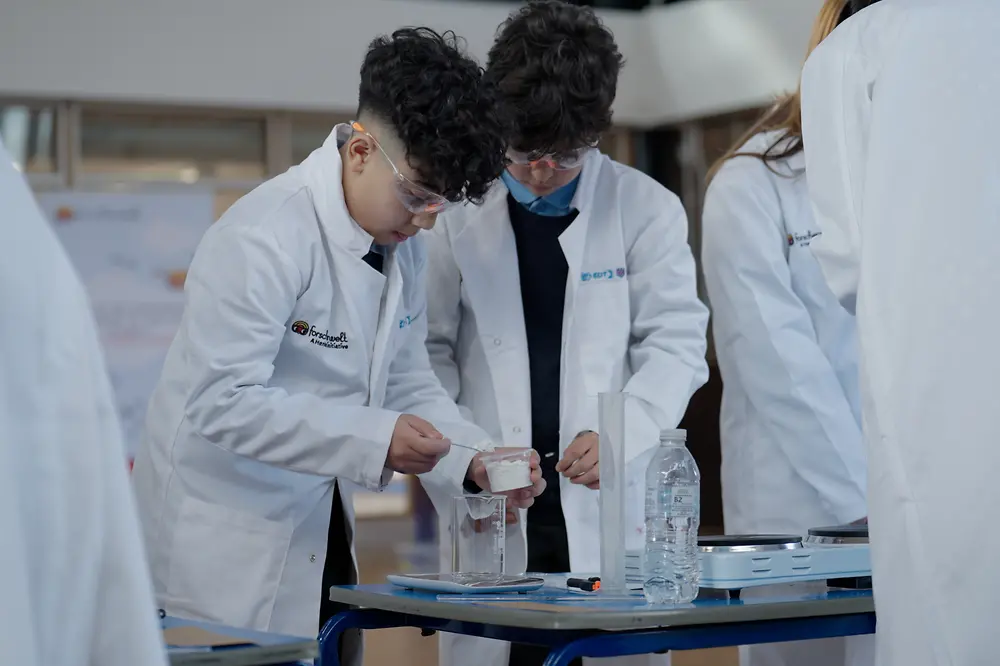 Two young boys wearing white lab coats and safety goggles conduct a science experiment.