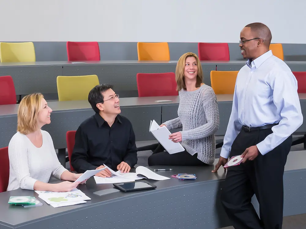 A standing employee is talking to three sitting employees.